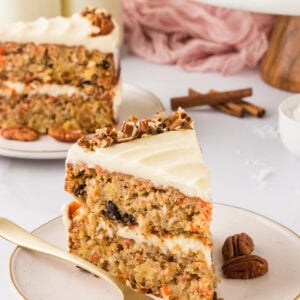 A close-up slice of gluten-free carrot cake on a plate with visible layers of cream cheese frosting, chopped pecans, and moist cake texture, surrounded by pecans and cinnamon sticks.