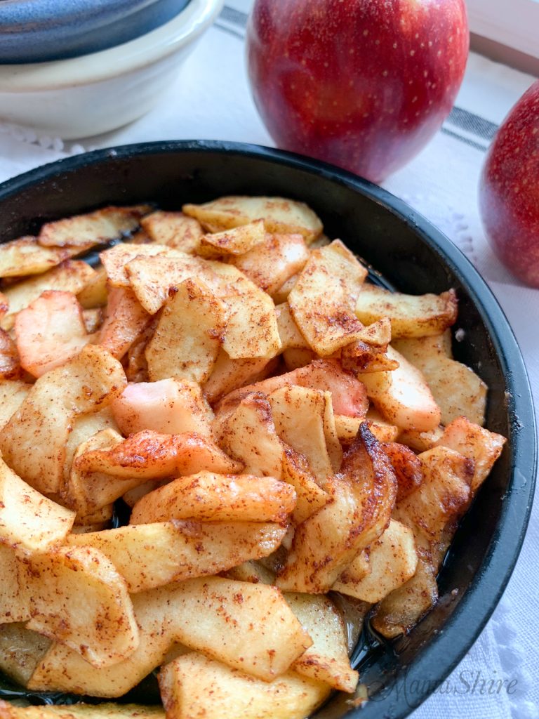 Air-fried spiced apples in a little black pan with a couple of red apples in the background.