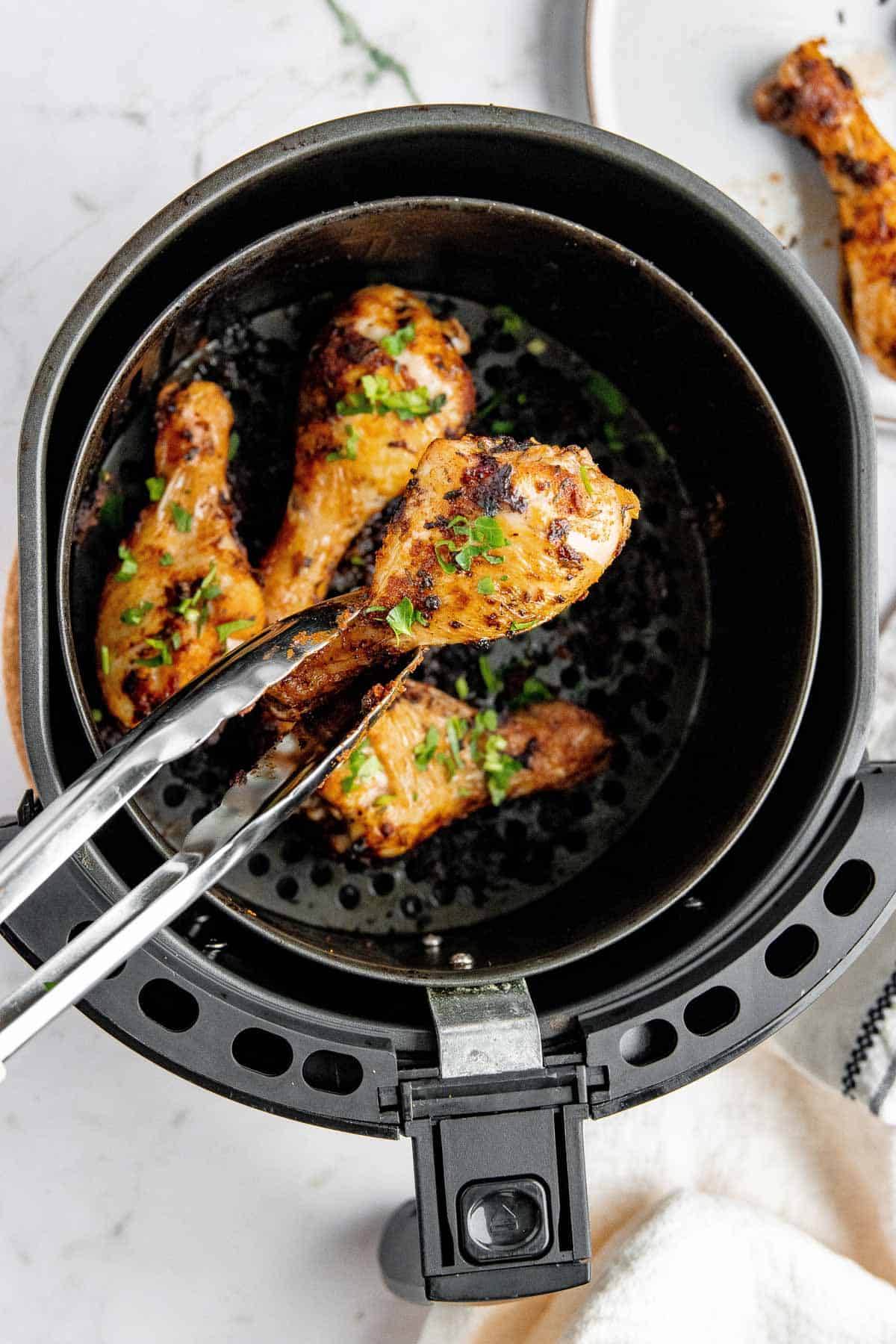 Cooking tongs picking up a chicken drum from an air fryer basket. 
