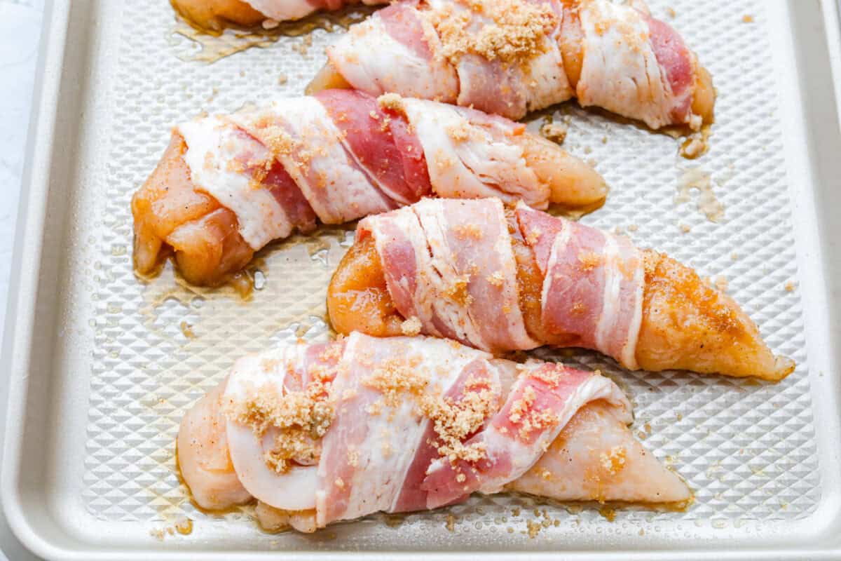 Bacon brown sugar chicken tenders are sweet, smoky, and slightly spicy. Made with only a few simple ingredients, they're like candied bacon hugging juicy chicken without being overly sweet. Great as a main or appetizer, and always a total hit! Bacon wrapped chicken tenders on a baking sheet with some more of the brown sugar mixture sprinkled on top.