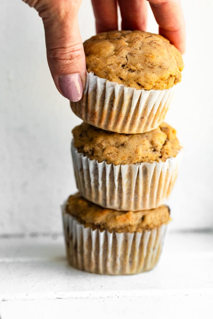 A hand stacking three freshly baked banana protein muffins in paper liners.