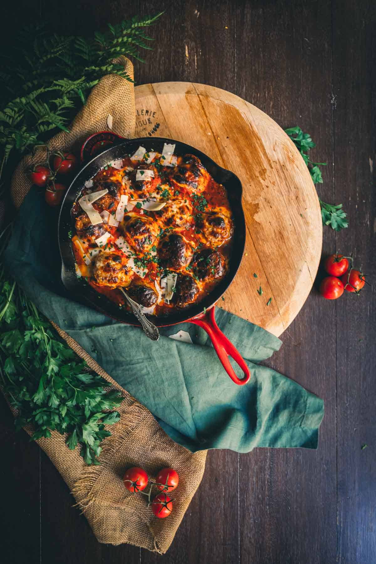 Overhead shot of a table with a skillet of chicken parmesan meatballs layered with melty cheese and fresh herbs ready to be served.