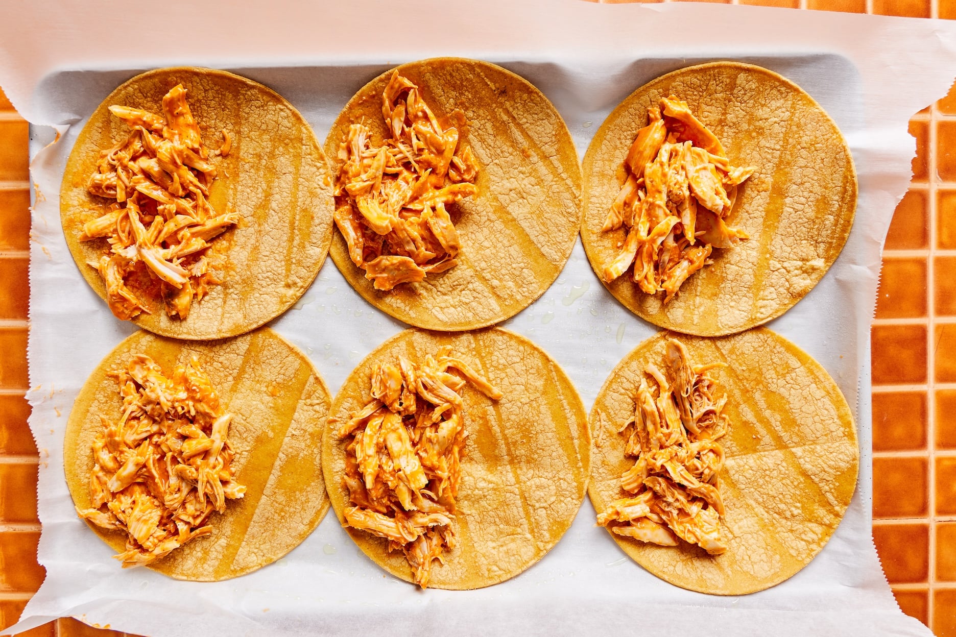Six tortillas laid out on a parchment-lined baking sheet with buffalo chicken spread out over one half of each tortilla. 