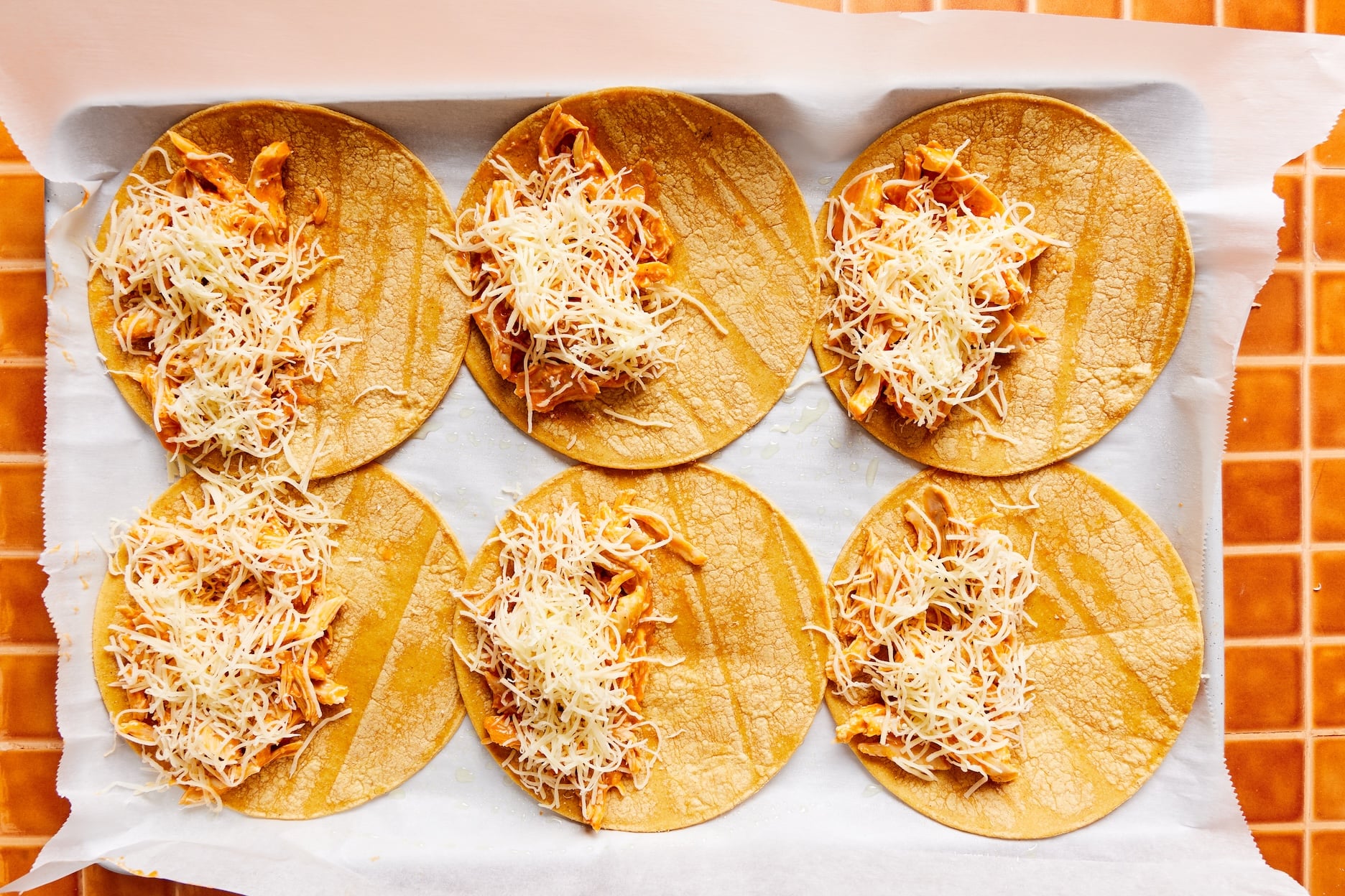 Six tortillas on a parchment-lined baking sheet with buffalo chicken and shredded cheese on one half of the tortilla. 