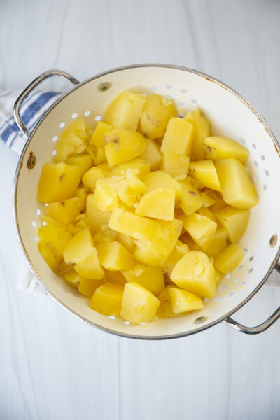 A white colander filled with peeled, chopped boiled potatoes sits on a light surface, ready for further preparation.