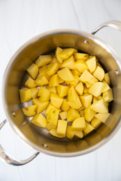 A stainless steel pot filled with evenly chopped yellow potatoes, ready for cooking, sits on a white countertop.