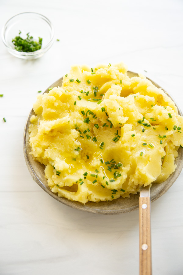A bowl of mashed potatoes garnished with chopped chives sits on a white surface, with a wooden-handled spoon and a small bowl of extra chives nearby.