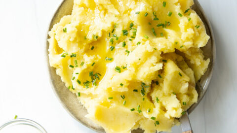 A plate of mashed potatoes topped with melted butter and chopped chives, with a small bowl of chives on the side. A wooden-handled spoon rests on the plate.