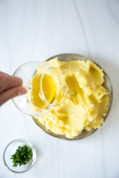 A hand pours melted butter from a small glass pitcher onto a bowl of mashed potatoes. A small bowl of chopped herbs sits nearby on a white surface.