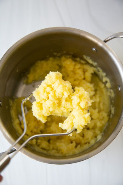 A stainless steel pot with mashed potatoes being lifted by a metal masher, showing a yellow, fluffy texture. The background is a bright white surface.