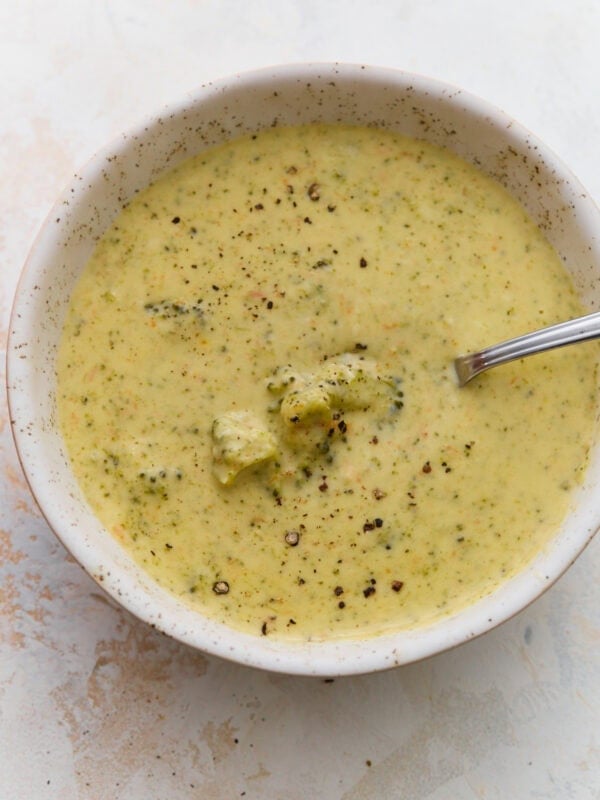 Bowl of gluten-free broccoli cheddar soup with a spoon resting in it, a whole broccoli floret in the center and cracked black pepper on top.