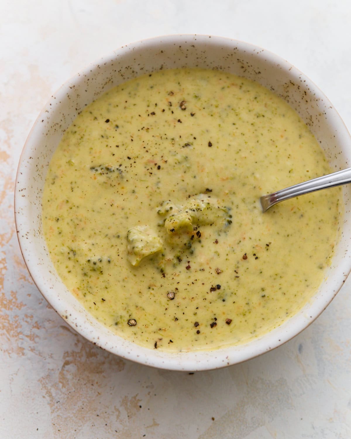 Bowl of gluten-free broccoli cheddar soup with a spoon resting in it, a whole broccoli floret in the center and cracked black pepper on top.