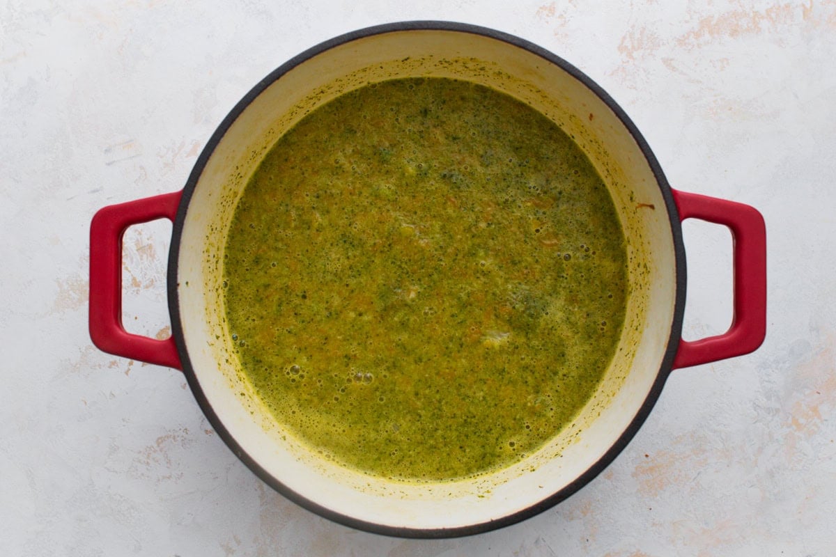 Broccoli, carrots, and broth simmered and partially blended into a chunky, bright green soup base in a red Dutch oven.