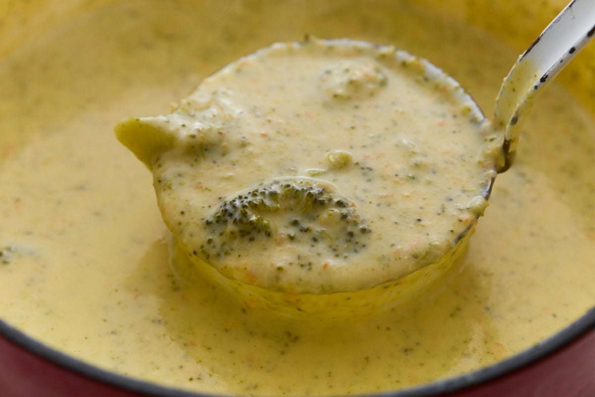 A ladle lifting a scoop of thick, creamy broccoli cheddar soup with a visible broccoli floret, straight from the pot.