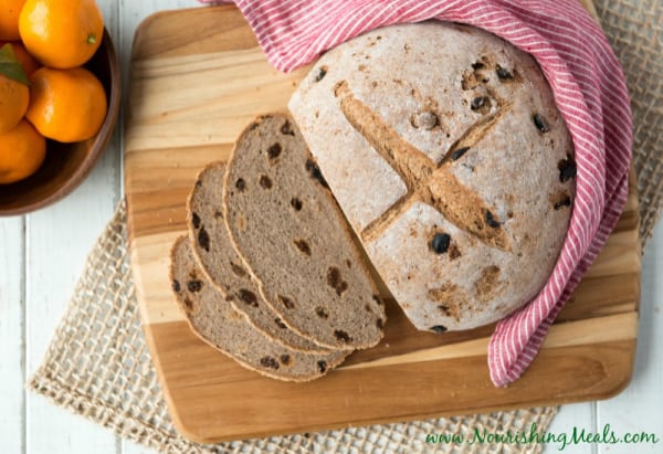 A round artisan loaf of Buckwheat Cinnamon Raisin Bread on a cutting board on a placement beside a red striped dish towel and bowl of clementines.