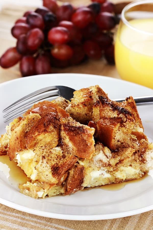 A serving of Gluten-Free Cinnamon Raisin Bread French Toast on white plate with fork, clump of red grapes, and glass of orange juice.