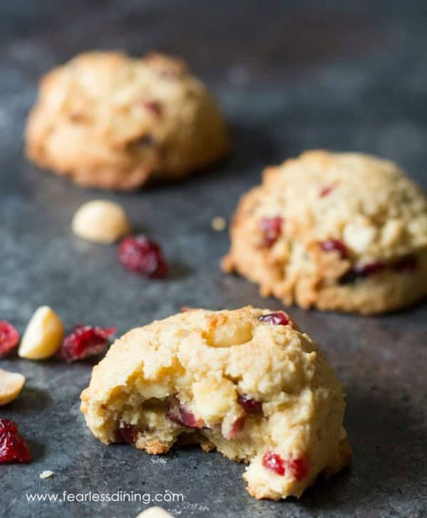 Gluten-Free Cranberry White Chocolate Macadamia Cookies from Fearless Dining on a slate surface beside macadamia nuts and cranberries. The cookie in front is half eaten.