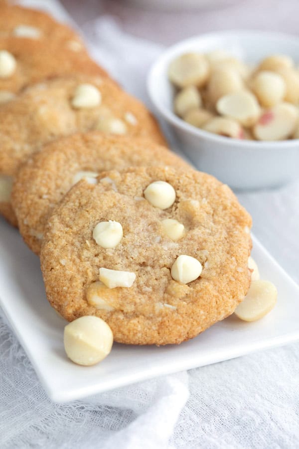 Gluten-Free and Keto White Chocolate Macadamia Nut Cookies from All Day I Dream About Food stacked domino stayle in a white rectangular dish beside a white saucer full of macadamia nuts on top of white gauze.