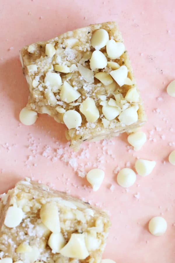 Two Gluten-Free No-Bake White Chocolate Macadamia Nut Bars from The Toasted Pine Nut on a pink tablecloth with white chocolate chips and sea salt scattered between them. 