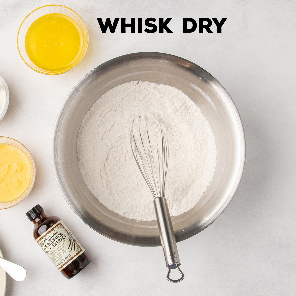 A whisk rests in a bowl of dry ingredients, with melted butter, beaten eggs, vanilla, and other ingredients arranged nearby. Text reads “WHISK DRY.”