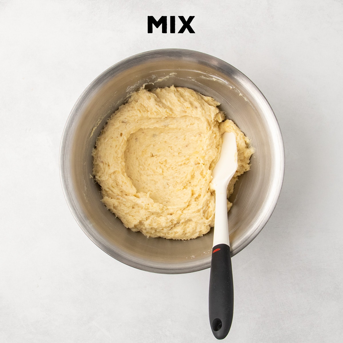 A thick pale yellow banana bread batter fills a metal mixing bowl with a spatula resting inside. Text reads “MIX.”