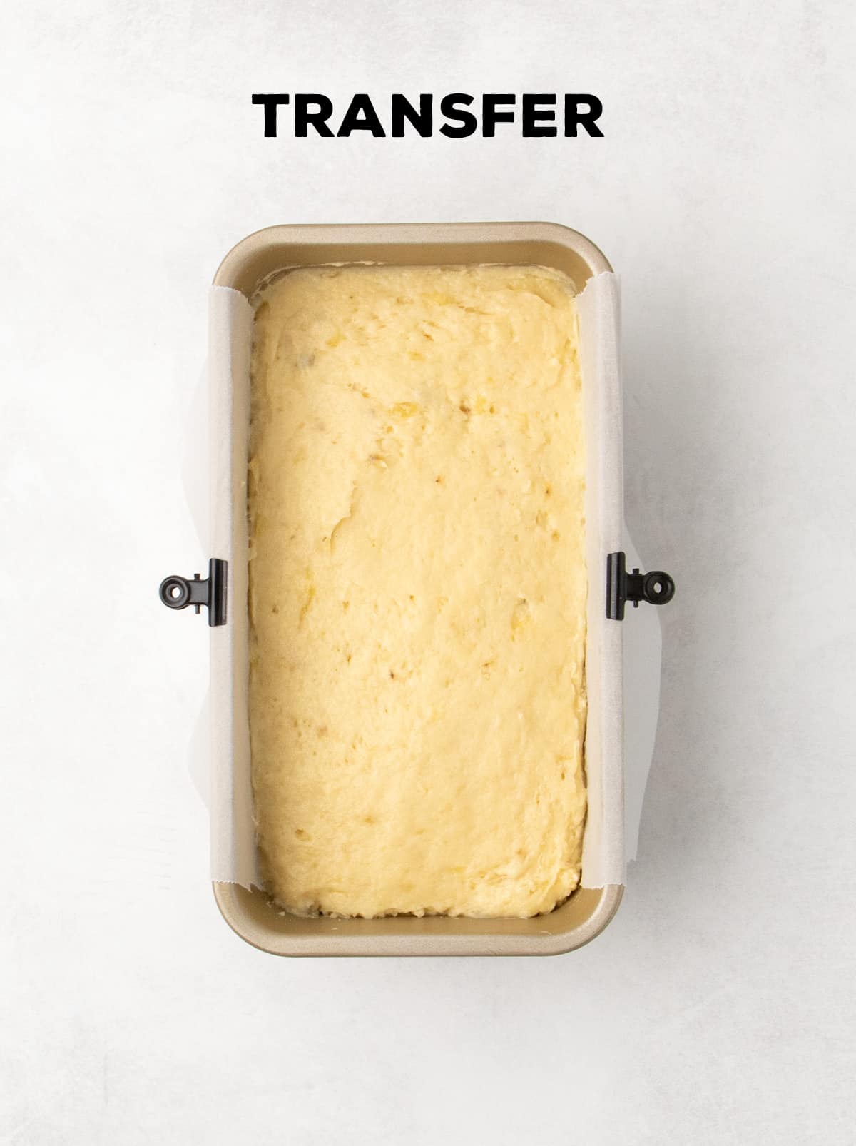 Raw light yellow banana bread batter is spread evenly in a parchment-lined loaf pan. Text reads “TRANSFER.”