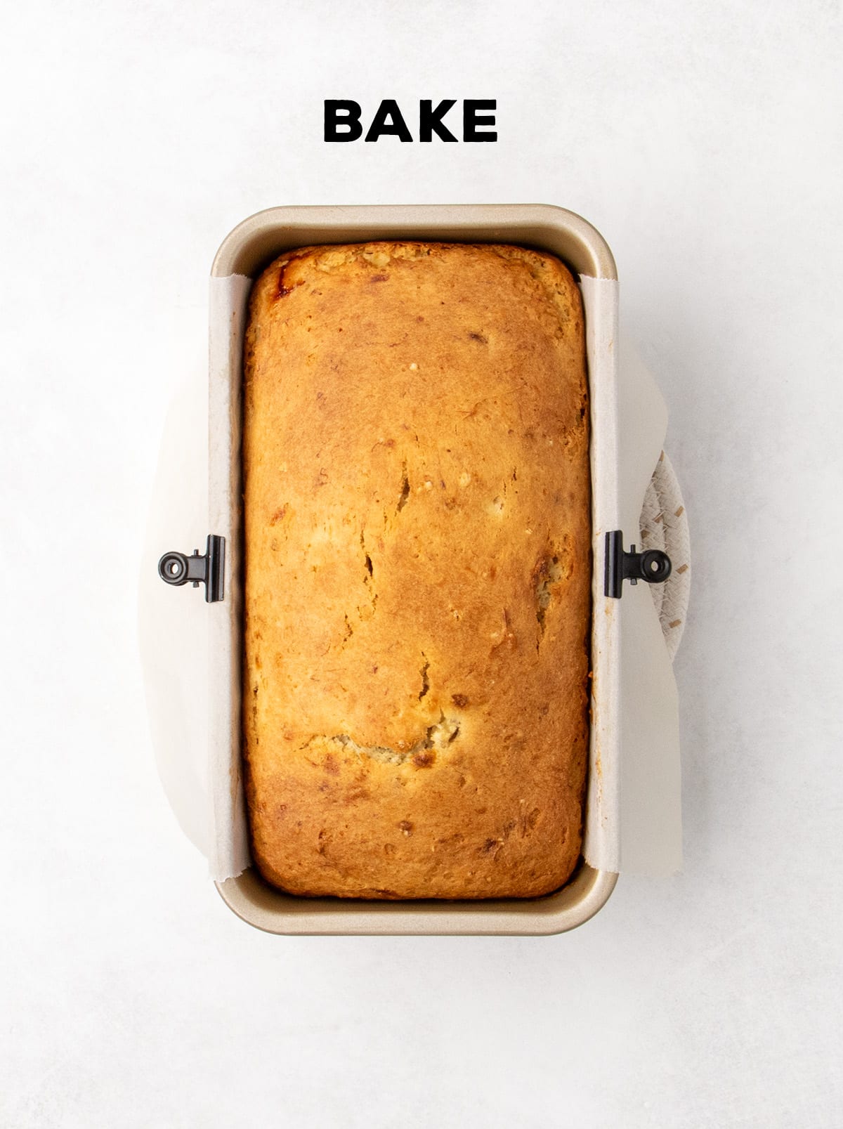 A baked loaf of banana bread sits in a parchment-lined loaf pan with a golden brown top. Text reads “BAKE.”
