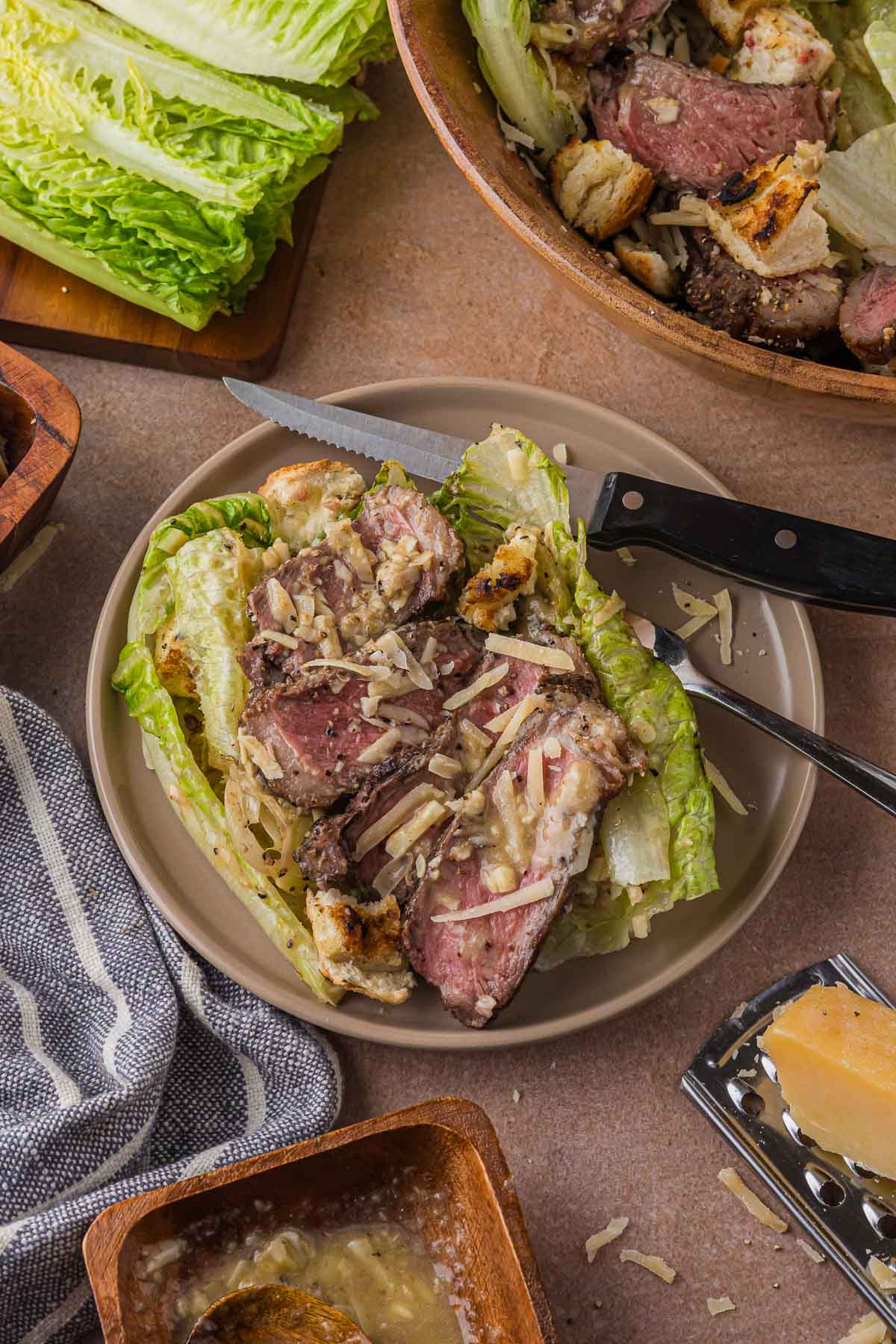 A plated serving of grilled steak Caesar salad beside the serving bowl, with a steak knife, raw romaine and a parmesan wedge on a grater nearby.