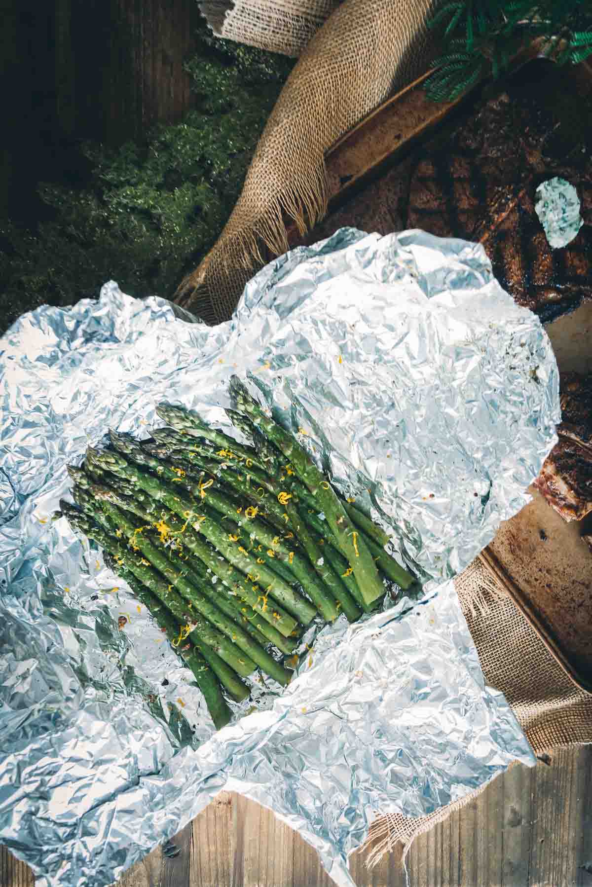 Unwrapped foil grilled asparagus on a table with steaks. 