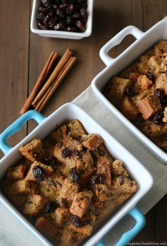 Two individual dishes containing Gluten-Free Cinnamon Raisin French Toast Casseroles beside three cinnamon sticks and bowl of raisins on hardwood surface.