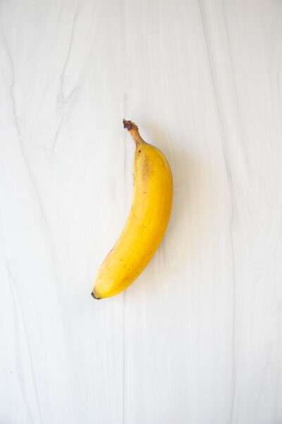 A single ripe yellow banana with a slightly brown stem is placed on a light-colored marble surface.