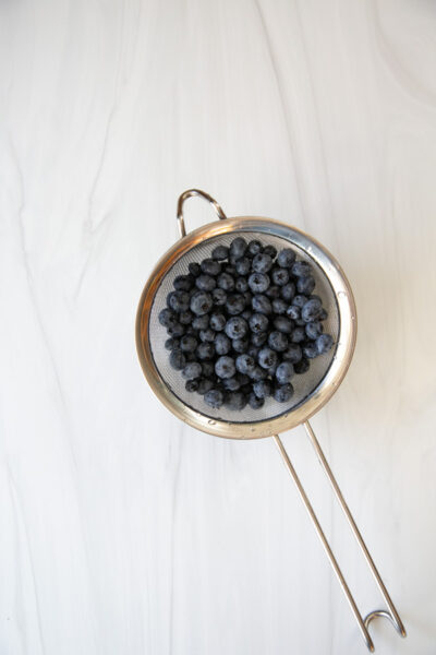 A metal strainer filled with fresh blueberries sits on a white marble surface.