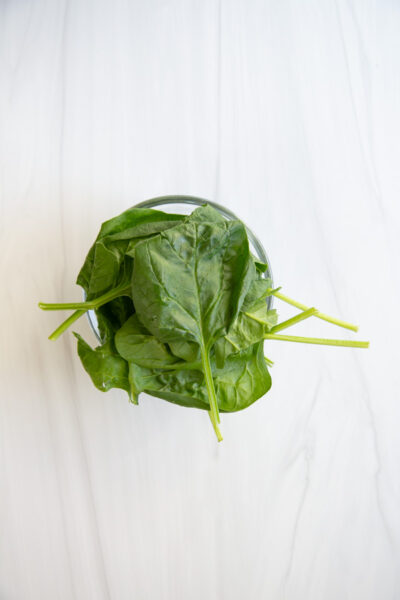 A small glass bowl filled with fresh spinach leaves, with some stems extending over the bowls edge, placed on a white marble surface.