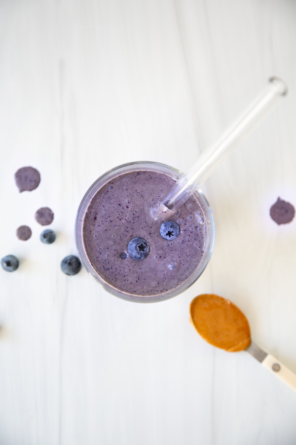 Overhead view of a glass of blueberry smoothie with a clear straw, topped with two blueberries. Nearby are scattered blueberries, smoothie drops, and a spoonful of peanut butter on a white surface.