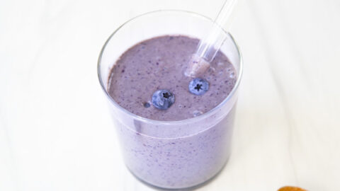 A glass of purple blueberry smoothie with a straw, topped with blueberries. Nearby, there are scattered blueberries and a spoon with peanut butter on a white surface.