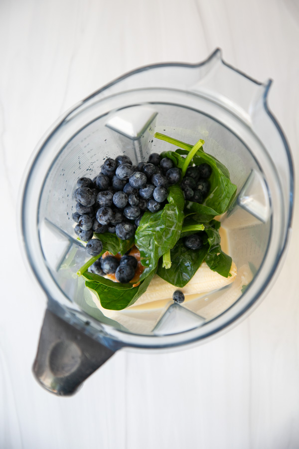 A blender containing fresh spinach leaves, blueberries, and a creamy liquid, viewed from above on a white surface.