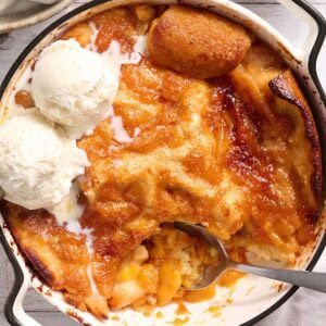 Gluten free peach cobbler in a white baking dish with three scoops of vanilla ice cream melting into the golden peach filling