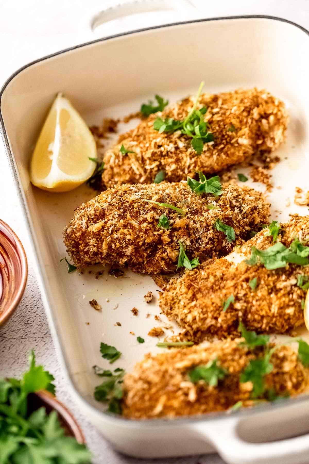 Parmesan panko breaded chicken breasts in a baking dish with lemon wedges and parsley.