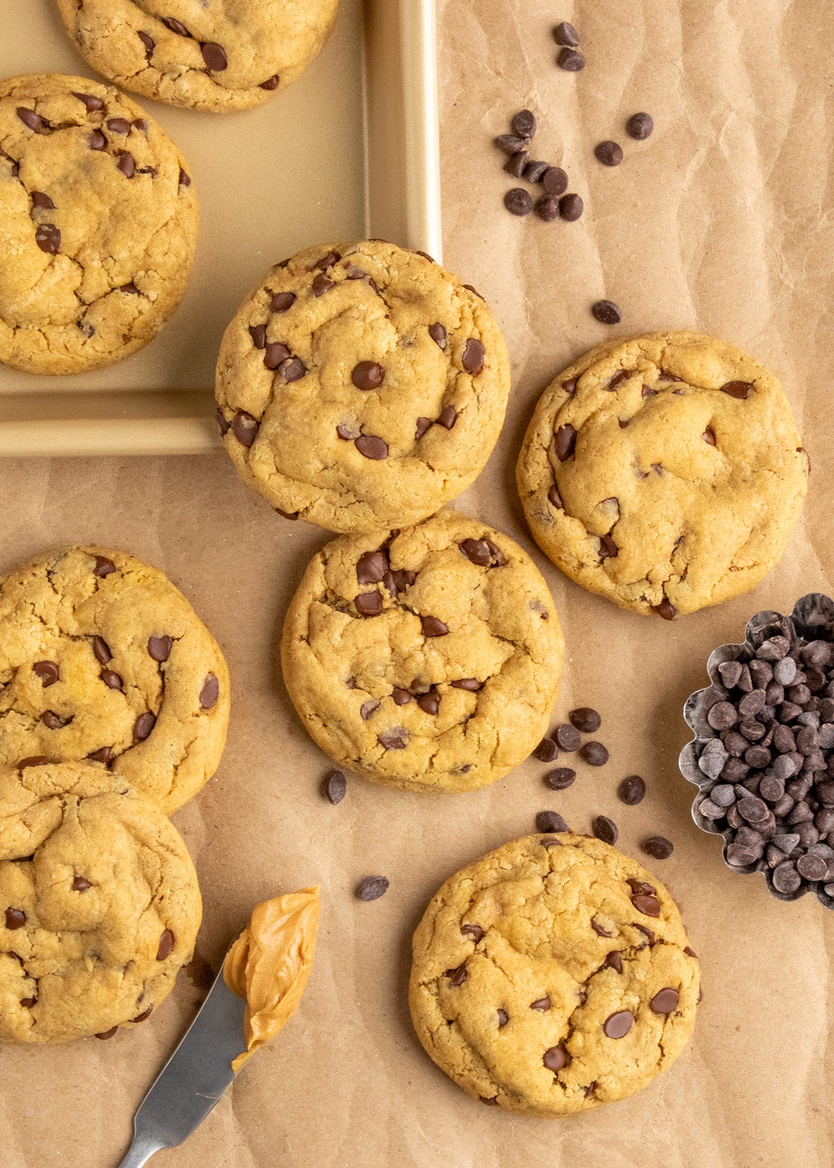 Peanut butter chocolate chip cookies are arranged on brown kraft paper with scattered chocolate chips, a dish of chocolate chips, and a knife holding a smear of peanut butter.