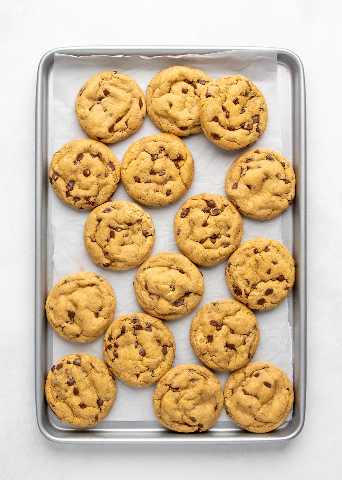 A parchment-lined baking sheet holds a 16 light brown round peanut butter cookies with mini chocolate chips, all viewed from above.