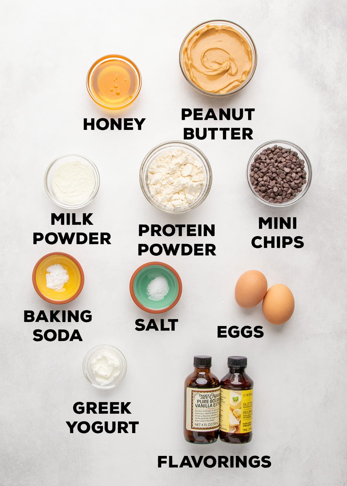 Bowls and containers hold honey, peanut butter, milk powder, protein powder, mini chocolate chips, baking soda, salt, Greek yogurt, eggs, and bottled flavorings, with each ingredient labeled.