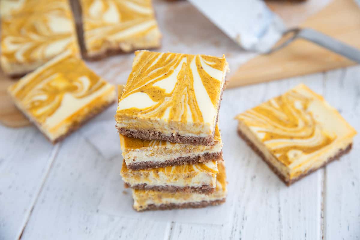 A stack of keto pumpkin swirl cheesecake bars on a white wooden table, in front of a cutting board with more bars.