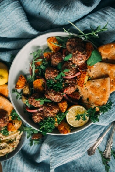 above shot of bowl filled with lamb meatballs, garnished with fresh herbs and lemon served with roasted potatoes and pita