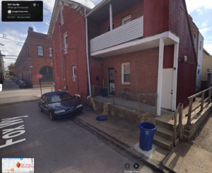 A google maps image of a red brick building with a small concrete porch and a foldingchair. Parked alongside is a blue sedan. It looks almost the same as the video of the ladies on the porch