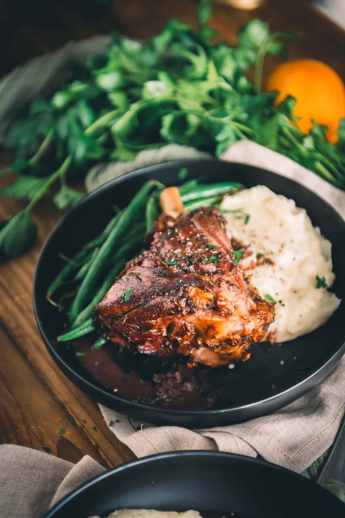 A plate with a sous vide lamb shank, mashed potatoes and green beans on a wooden table.
