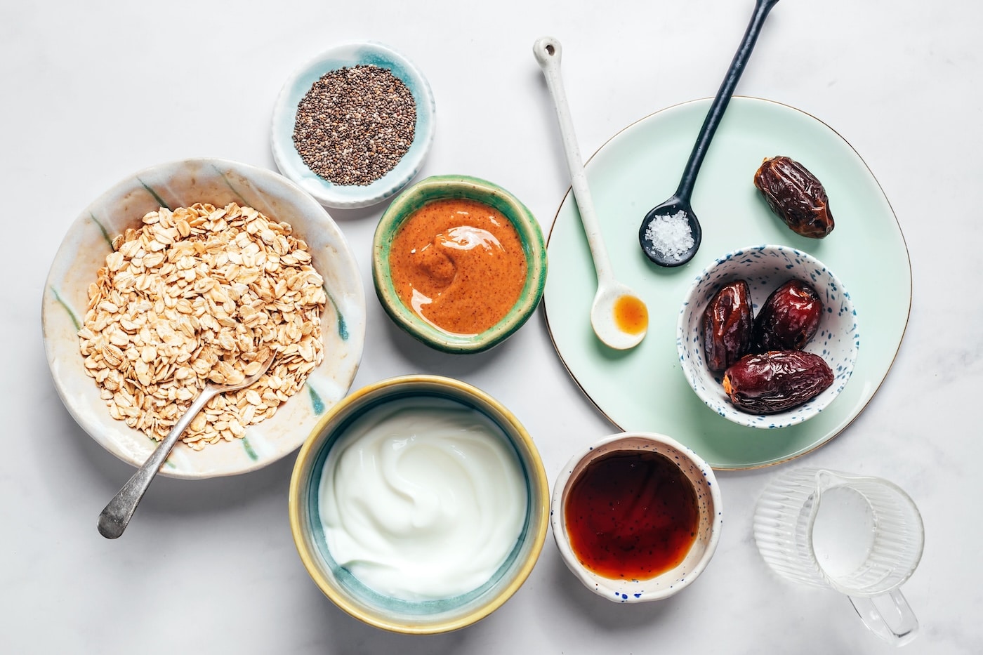Rolled oats, dates, chia seeds, yogurt, nut butter, and maple syrup arranged on a white surface.