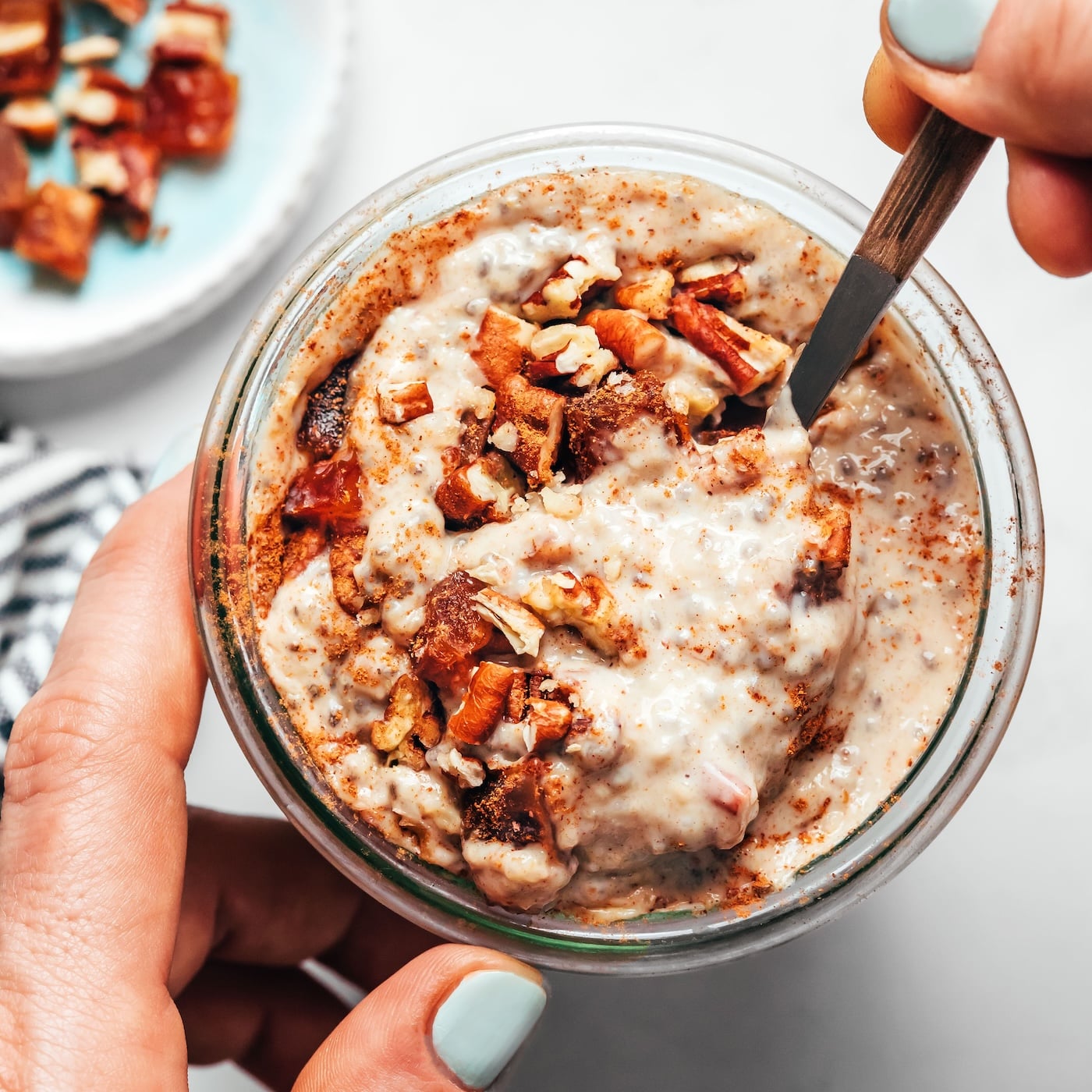 A woman’s hands hold and stir a jar of overnight oats topped with chopped dates, pecans, and cinnamon.