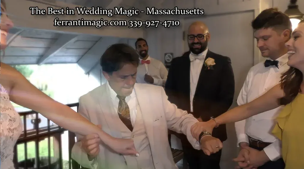 WeddingMagicianMassachusetts The Magic of Joe Ferranti The Magic of Joe Ferranti Wedding party in formal attire watches a man in a cream suit perform a magic trick as guests link arms nearby in a corridor
