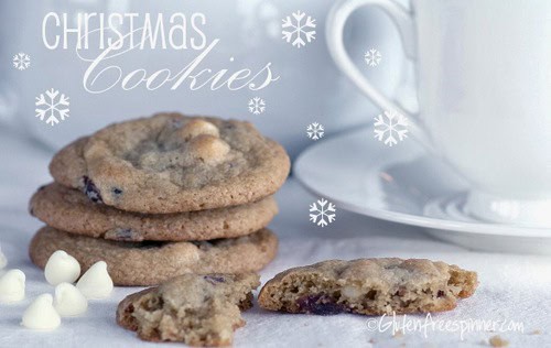 Gluten-Free White Chocolate and Cherry Macadamia Cookies from Gluten-Free Spinner on a white cloth with one broken in half in front and a stack of three behind it. White chocolate chips, a white cup and saucer, and a background of snowflakes complete the photo.