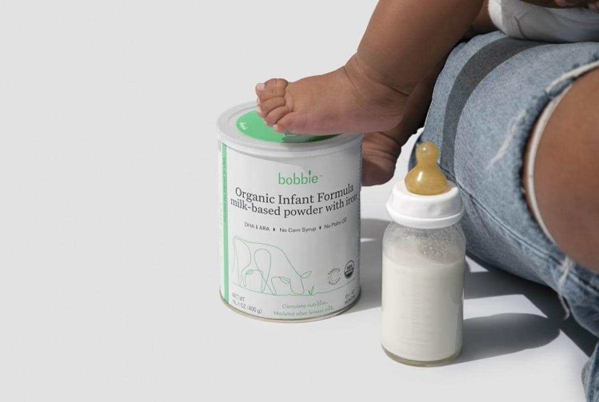 A can of bobbie formula and a small baby bottle next to a baby foot and parent leg.